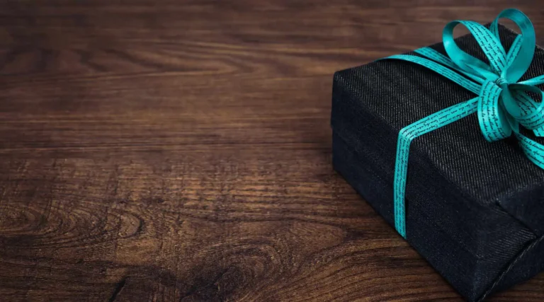 Nell'immagine, il primo piano su un pacco regalo con fiocco azzurro poggiato su un tavolo di legno.